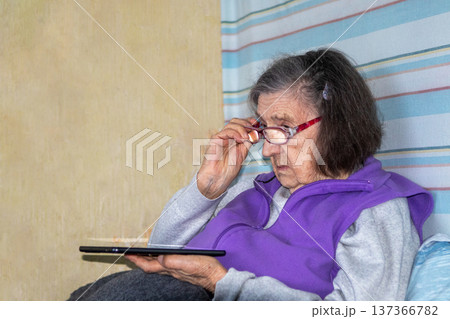 Elderly woman reading on a tablet with glasses at home, concentrating while seated Elderly woman reading on a tablet with glasses at home, concentrating while seated 137366782