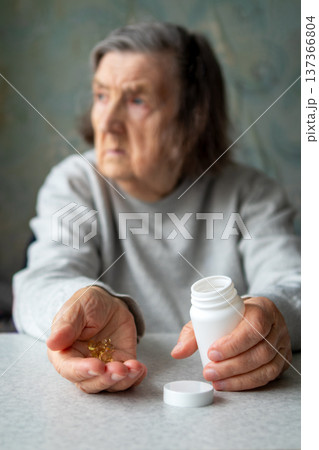 Elderly woman holding medication bottle and pills at home showing concern and vulnerability 137366804