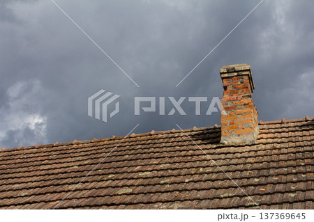 Cloudy sky over a traditional brick chimney on a rustic roof in late afternoon light highlighting the weathered tiles and contrasting textures Cloudy sky over a traditional brick chimney on a rustic roof in late afternoon light highlighting the weathered tiles and contrasting textures 137369645
