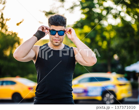 Fit man wearing sunglasses and black tank top standing outdoors in city park Fit man wearing sunglasses and black tank top standing outdoors in city park 137371010