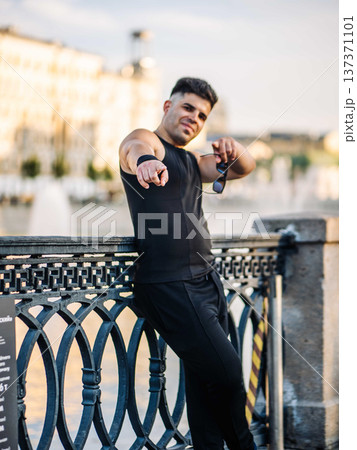 Young man pointing at viewer, standing by urban bridge railing in a city Young man pointing at viewer, standing by urban bridge railing in a city 137371101