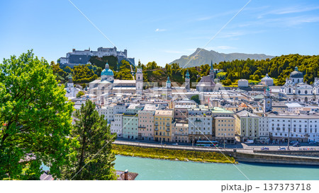 The charming city of Salzburg, Austria, showcases colorful buildings along the Danube River with a historic castle perched atop a hill, set against a clear blue sky. 137373718