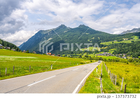 A road winds through green fields and mountains in the Alps. Small houses are visible along the route under a cloudy sky. This scene captures the beauty of nature and rural life. 137373732