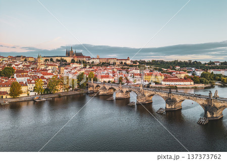 A serene morning scene captures Prague Castle rising above the city, with Charles Bridge stretching over the Vltava River. The old town awakens with soft light and vibrant rooftops. 137373762