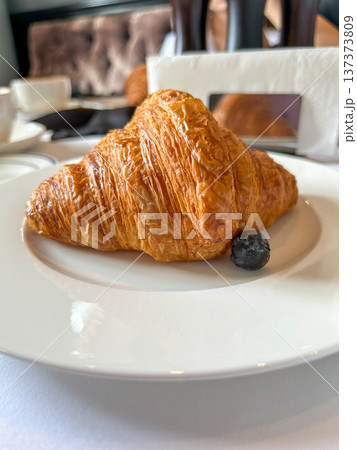 Fresh golden croissant on a white plate with a blueberry, close-up breakfast pastry on a restaurant table Fresh golden croissant on a white plate with a blueberry, close-up breakfast pastry on a restaurant table 137373809