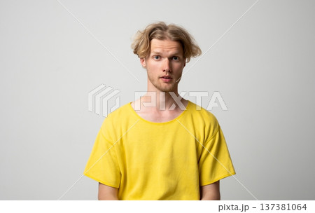 Young man standing with a neutral expression, showing surprise or inquiry, wearing a bright yellow t-shirt and maintaining eye contact on a plain background 137381064