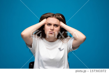 Stressed young woman enduring headache and pressure, showing anxiety and frustration on blue background Stressed young woman enduring headache and pressure, showing anxiety and frustration on blue background 137381072