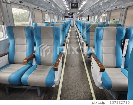 Interior view of a modern clean train carriage with comfortable blue and grey ergonomic seating, featuring bright fluorescent lighting, overhead monitors, and large windows showing green scenery. 137382759