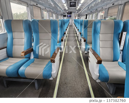 Interior view of a modern clean train carriage with comfortable blue and grey ergonomic seating, featuring bright fluorescent lighting, overhead monitors, and large windows showing green scenery. Interior view of a modern clean train carriage with comfortable blue and grey ergonomic seating, featuring bright fluorescent lighting, overhead monitors, and large windows showing green scenery. 137382760