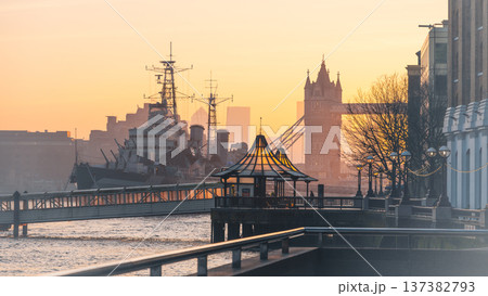 Morning light casts silhouettes of HMS Belfast and Tower Bridge along the River Thames. People stroll near the water as the day begins in London, UK. Morning light casts silhouettes of HMS Belfast and Tower Bridge along the River Thames. People stroll near the water as the day begins in London, UK. 137382793