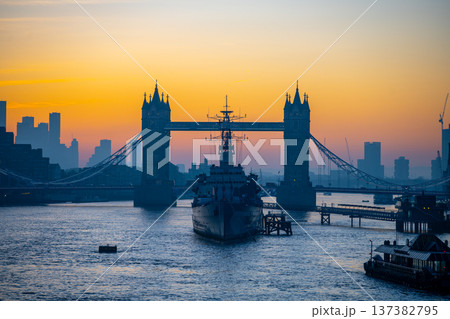 At dawn, the river Thames shows the silhouette of Tower Bridge and HMS Belfast. The scene captures the urban landscape of London as the sun rises over the city. At dawn, the river Thames shows the silhouette of Tower Bridge and HMS Belfast. The scene captures the urban landscape of London as the sun rises over the city. 137382795