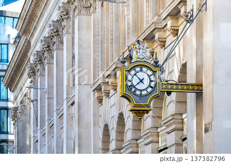 A street clock hangs on the Bank building in the City of London. The clock shows the time and adds character to the historic architecture. People walk by in this busy area of the city. 137382796