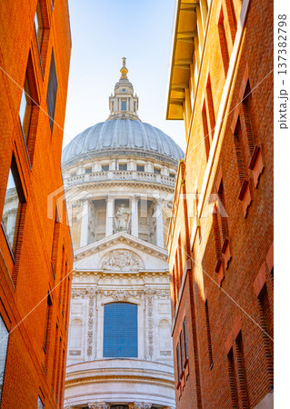 Visitors walk along a narrow street in London, looking up at St Pauls Cathedral. The sunlight highlights the dome and architectural details of the building. 137382798
