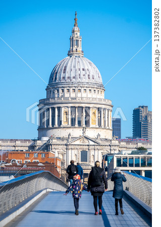 People walk across Millennium Bridge towards St Pauls Cathedral in London. The dome of the cathedral stands prominently against a clear blue sky. People walk across Millennium Bridge towards St Pauls Cathedral in London. The dome of the cathedral stands prominently against a clear blue sky. 137382802