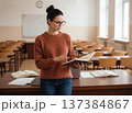 Focused young female teacher in glasses reading a book in an empty classroom setting, copy space. 137384867
