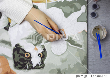 View from above table of artist hand creating animal themed piece of art. Cat is drawn on canvas by girl artist. Concentration on drawing and painting. Girl holding brush with paint in her hand 137386138