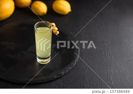 Tall shot glass of limoncello garnished with a curled lemon peel, surrounded by fresh lemons on dark textured background 137386489