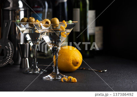 Two martini glasses with skewered green olives and citrus twists against dark surface with wine bottles blurred in the background Two martini glasses with skewered green olives and citrus twists against dark surface with wine bottles blurred in the background 137386492