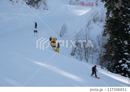 Snowboarder descending snowy slope with snowmaking machine and winter mountain landscape at Krasnaya Polyana ski resort in Sochi region. Winter tourism marketing, ski resort promotion, outdoor sport Snowboarder descending snowy slope with snowmaking machine and winter mountain landscape at Krasnaya Polyana ski resort in Sochi region. Winter tourism marketing, ski resort promotion, outdoor sport 137386651