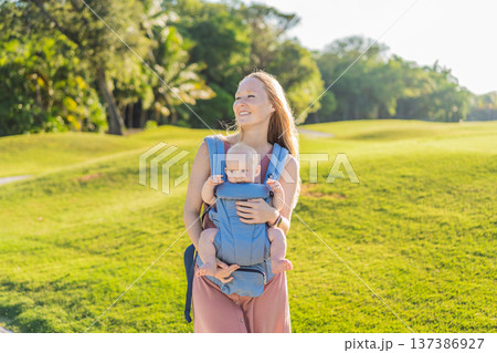 Mother using ergonomic baby carrier during a walk with her infant outdoors. Comfortable babywearing, active motherhood lifestyle, bonding, modern parenting, hands free care and everyday family routine Mother using ergonomic baby carrier during a walk with her infant outdoors. Comfortable babywearing, active motherhood lifestyle, bonding, modern parenting, hands free care and everyday family routine 137386927