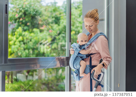 Woman vacuuming a room while carrying her baby in an ergonomic carrier. Modern motherhood, multitasking parent, home chores with infant, balance of childcare and housework concept. Woman vacuuming a room while carrying her baby in an ergonomic carrier. Modern motherhood, multitasking parent, home chores with infant, balance of childcare and housework concept. 137387051