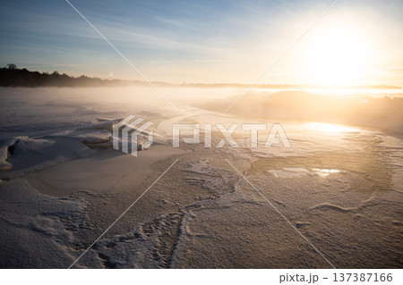 Landscape of frozen lake covered in snow and ice in winter season, sun rays beam through misty air. 137387166