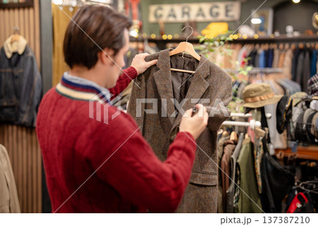 Man hipster inspecting quality of trendy used jacket in fashionable men's clothing vintage store. Man hipster inspecting quality of trendy used jacket in fashionable men's clothing vintage store. 137387210