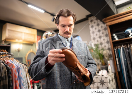 Serious handsome man customer thinking on buying fancy leather shoes in second hand vintage shop. Serious handsome man customer thinking on buying fancy leather shoes in second hand vintage shop. 137387215