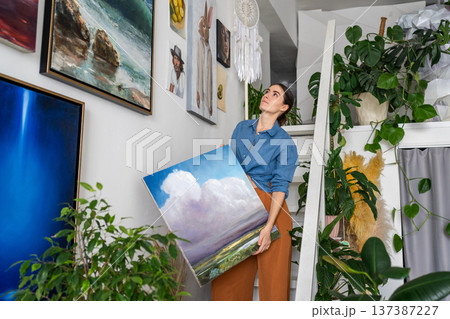 Pleased confident woman artist stands on ladder in loft studio with new artwork in art workshop. Pleased confident woman artist stands on ladder in loft studio with new artwork in art workshop. 137387227