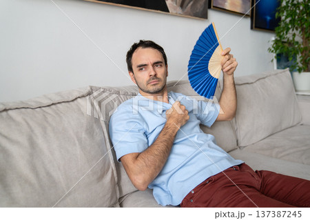 Sad man suffering from heat in apartment, fanning himself with paper fan, no air conditioner at home Sad man suffering from heat in apartment, fanning himself with paper fan, no air conditioner at home 137387245