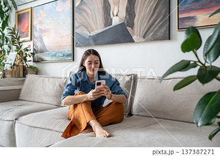 Curious woman artist sitting on couch watching media online on smartphone at cozy home gallery. 137387271