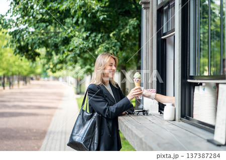 Happy cheerful businesswoman buying ice cream from street kiosk in city park after busy work day. Happy cheerful businesswoman buying ice cream from street kiosk in city park after busy work day. 137387284