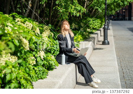 Relaxed businesswoman on daily break in park sitting under trees smiles and squints in bright sun. Relaxed businesswoman on daily break in park sitting under trees smiles and squints in bright sun. 137387292