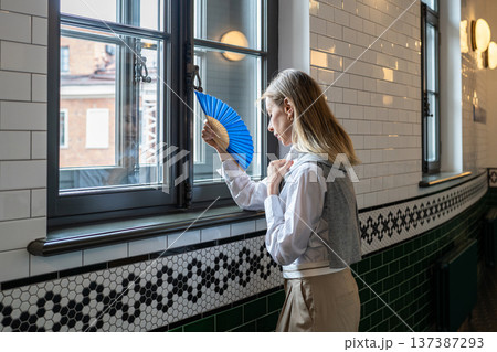 Woman feeling suffering from heatstroke and dizziness indoors, waving paper fan in hand to cool down Woman feeling suffering from heatstroke and dizziness indoors, waving paper fan in hand to cool down 137387293
