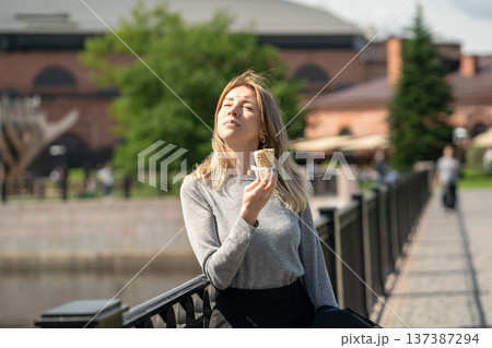 Carefree business woman enjoys sunny weather and ice cream, stands on bridge squinting at bright sun 137387294