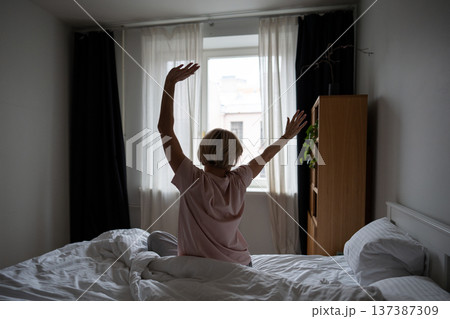 Rested positive woman sitting in bed in bedroom, stretching arms and body after good night sleep. 137387309
