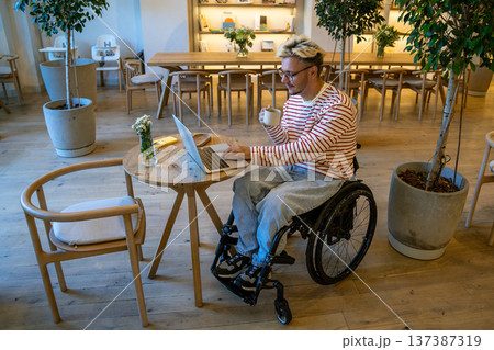 Cheerful positive disabled man in wheelchair working on laptop and drinking coffee in cozy cafe. Cheerful positive disabled man in wheelchair working on laptop and drinking coffee in cozy cafe. 137387319
