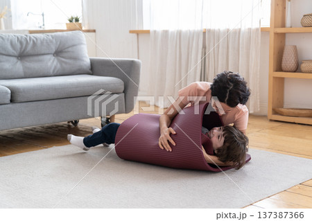 Mother wrapped smiling kid in yoga mat playing with son after sport exercise at home during lockdown Mother wrapped smiling kid in yoga mat playing with son after sport exercise at home during lockdown 137387366