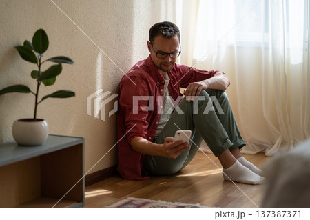 Upset young man holding smartphone waiting for ex-girlfriend call while sitting on floor at home Upset young man holding smartphone waiting for ex-girlfriend call while sitting on floor at home 137387371