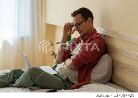 Concentrated Caucasian man sits on bed in hotel room reading news via internet or checking email Concentrated Caucasian man sits on bed in hotel room reading news via internet or checking email 137387429