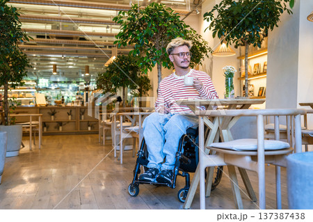 Relaxed positive disabled man in wheelchair drinking coffee in trendy cafe, staying socially active. 137387438