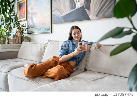 Happy relaxed woman artist sitting on couch at loft gallery apartment texting on smartphone online. Happy relaxed woman artist sitting on couch at loft gallery apartment texting on smartphone online. 137387479