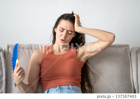 Stressed tired woman suffering from summer heat at home, sitting on sofa waving paper fan. Stressed tired woman suffering from summer heat at home, sitting on sofa waving paper fan. 137387483