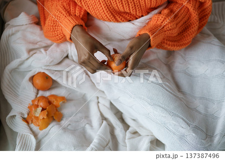 African woman hands peeling ripe sweet tangerine, wear orange sweater, lying in bed under the plaid 137387496