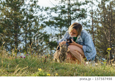 Woman photographer plays with lonely cat walking on forest meadow against blurry green trees 137387498