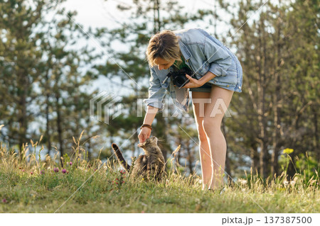 Woman photographer plays with lonely cat walking on forest meadow against blurry green trees Woman photographer plays with lonely cat walking on forest meadow against blurry green trees 137387500