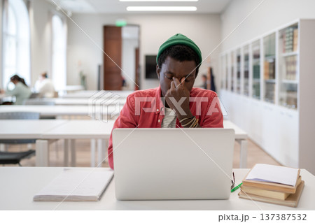 Tired black student guy massaging nose sitting with closed eyes in front of laptop in library Tired black student guy massaging nose sitting with closed eyes in front of laptop in library 137387532