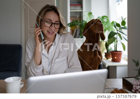 Woman in pajamas sitting with dog in living room, taking on mobile phone, remote working at laptop Woman in pajamas sitting with dog in living room, taking on mobile phone, remote working at laptop 137387679