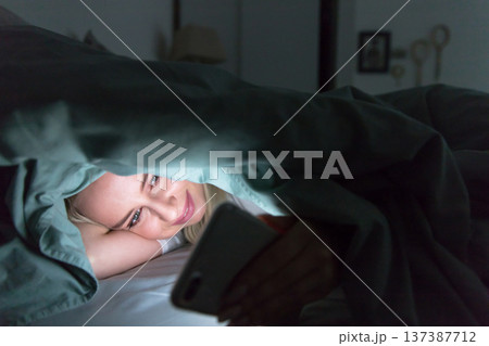 Woman lying in bed under blanket using smartphone at night.Insomnia, melatonin, internet addiction Woman lying in bed under blanket using smartphone at night.Insomnia, melatonin, internet addiction 137387712
