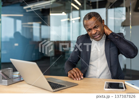 Professional african american man experiencing intense neck and shoulder discomfort while working on a laptop in a modern office, struggling with work-related stress and poor posture 137387826
